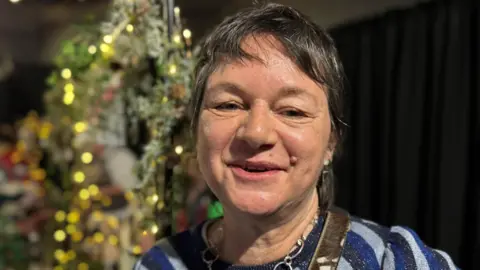 امرأة ذات شعر داكن قصير تشير بالكاميرا. وهناك لافتات مضيئة خلفها