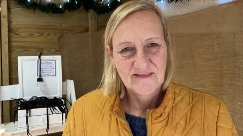 صورة لامرأة ترتدي سترة صفراء داكنة وتباع منتجات صواني القطط خلفها وعود عيد الميلاد وأضواء عيد الميلاد فوق رأسها